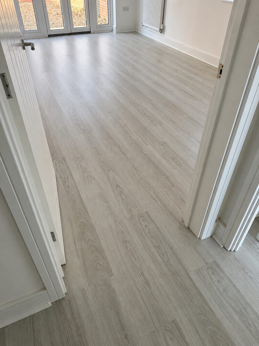 Pale grey laminate flooring running between a hallway and a bright room with French doors